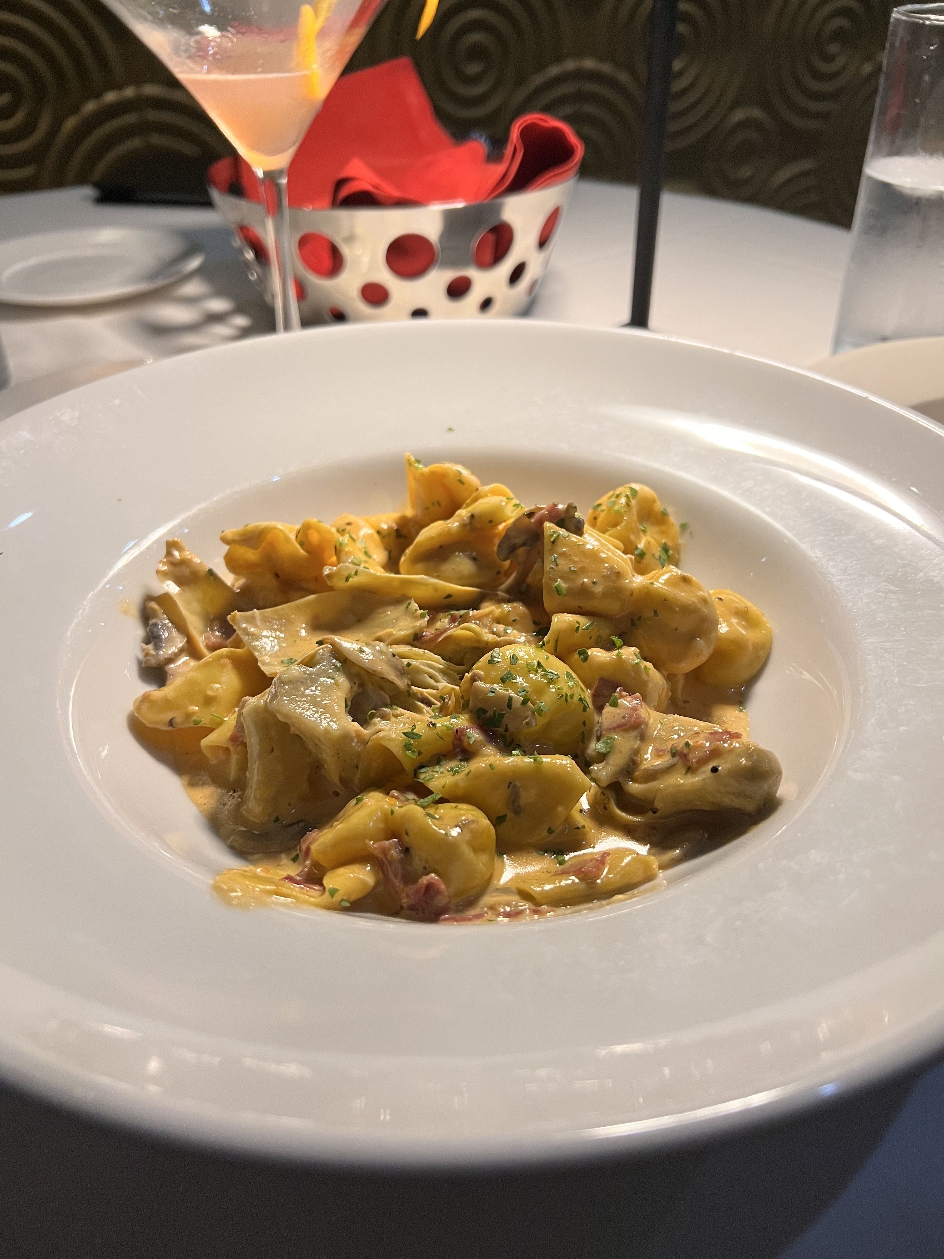 A close-up of a creamy tortellini pasta dish garnished with herbs, served in a white bowl on a restaurant table with a cocktail, bread basket, and water glass in the background.