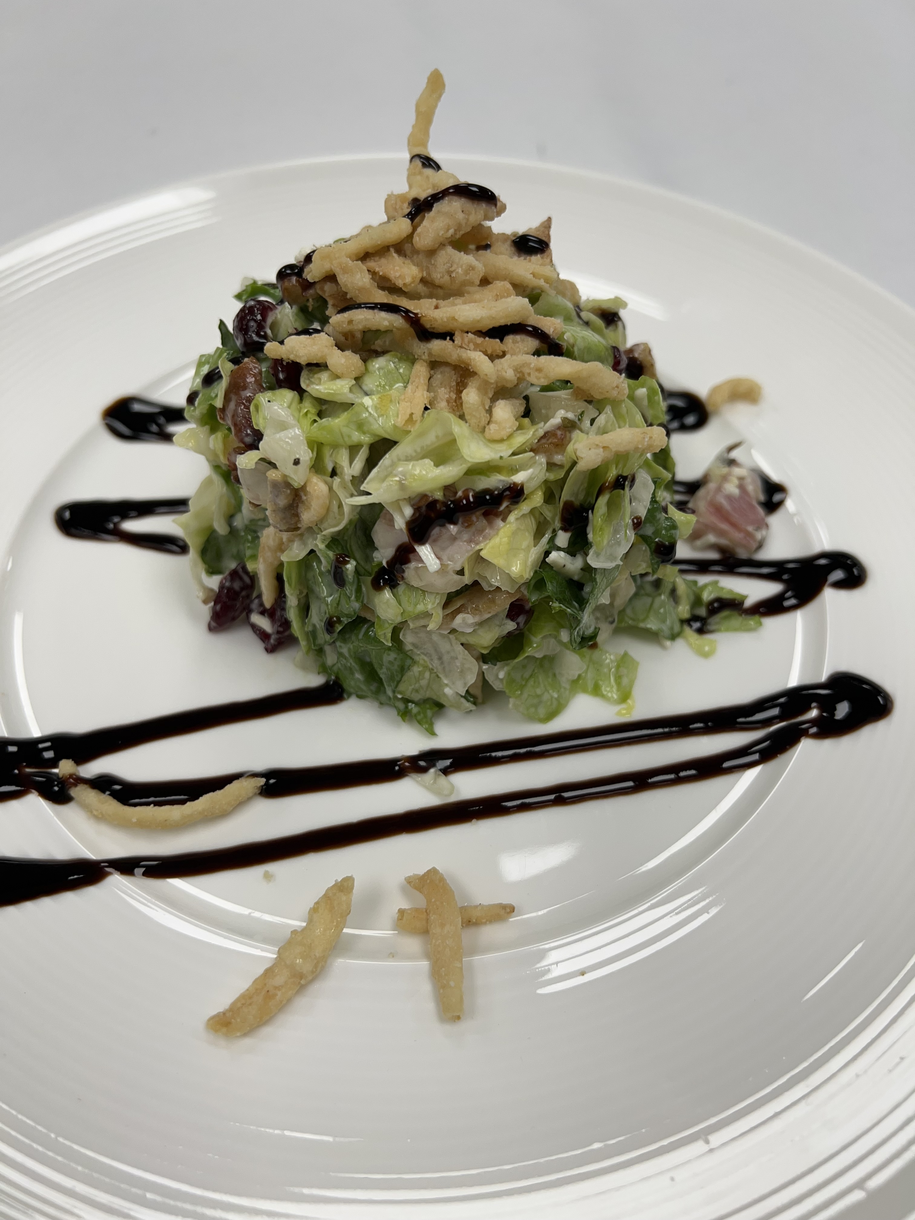 A neatly stacked chopped salad made with crisp romaine lettuce, dried cranberries, and bits of bacon, topped with crunchy fried onions and a drizzle of dark balsamic glaze. The salad is plated on a white dish with artistic lines of balsamic glaze and a few scattered fried onion pieces for garnish.
