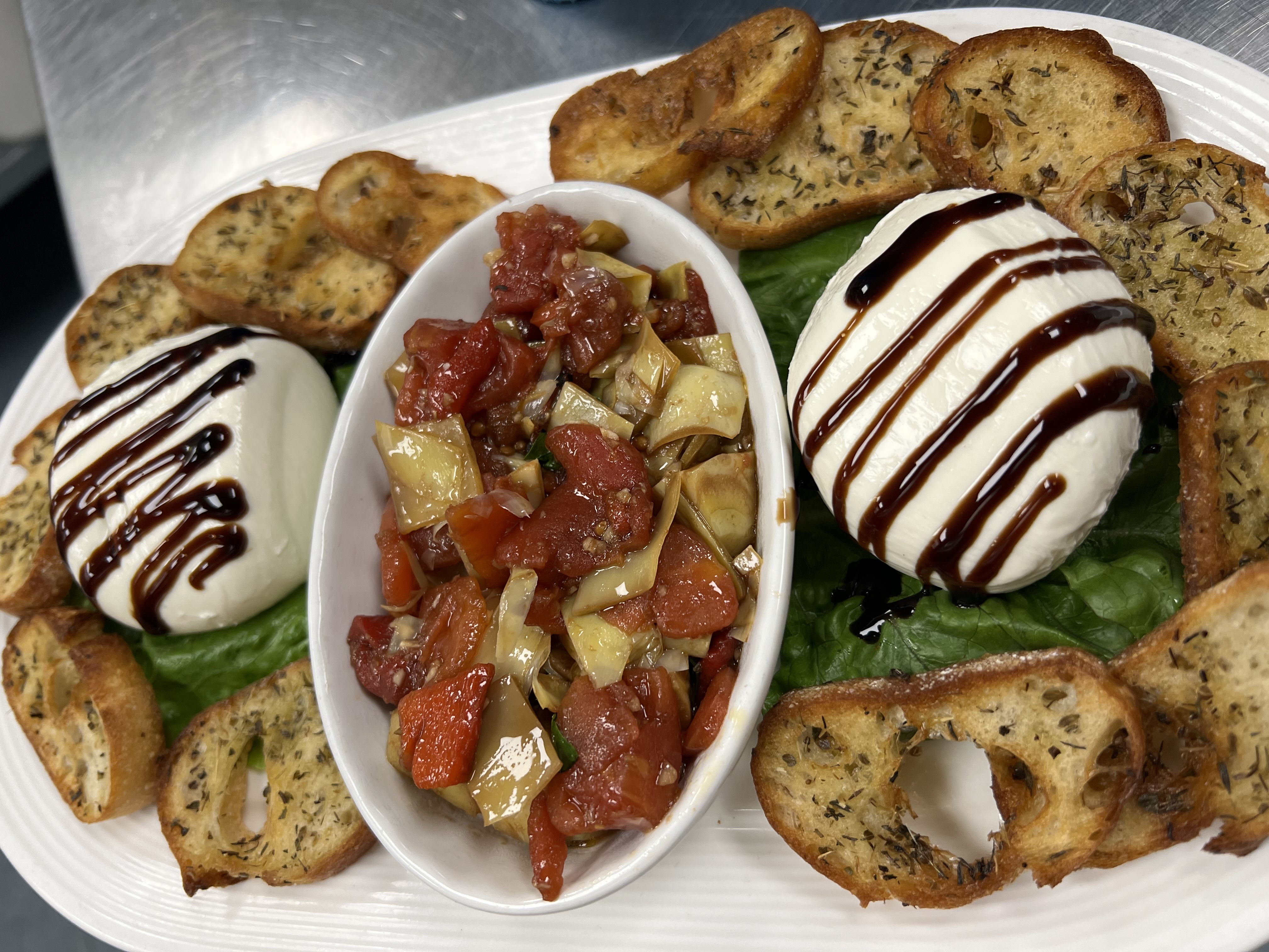 A beautifully plated appetizer featuring two balls of fresh mozzarella drizzled with balsamic glaze, surrounded by crispy herb crostini on a bed of green leaf lettuce. In the center is a small dish filled with a colorful tomato and artichoke bruschetta mix, creating a vibrant and appetizing presentation.