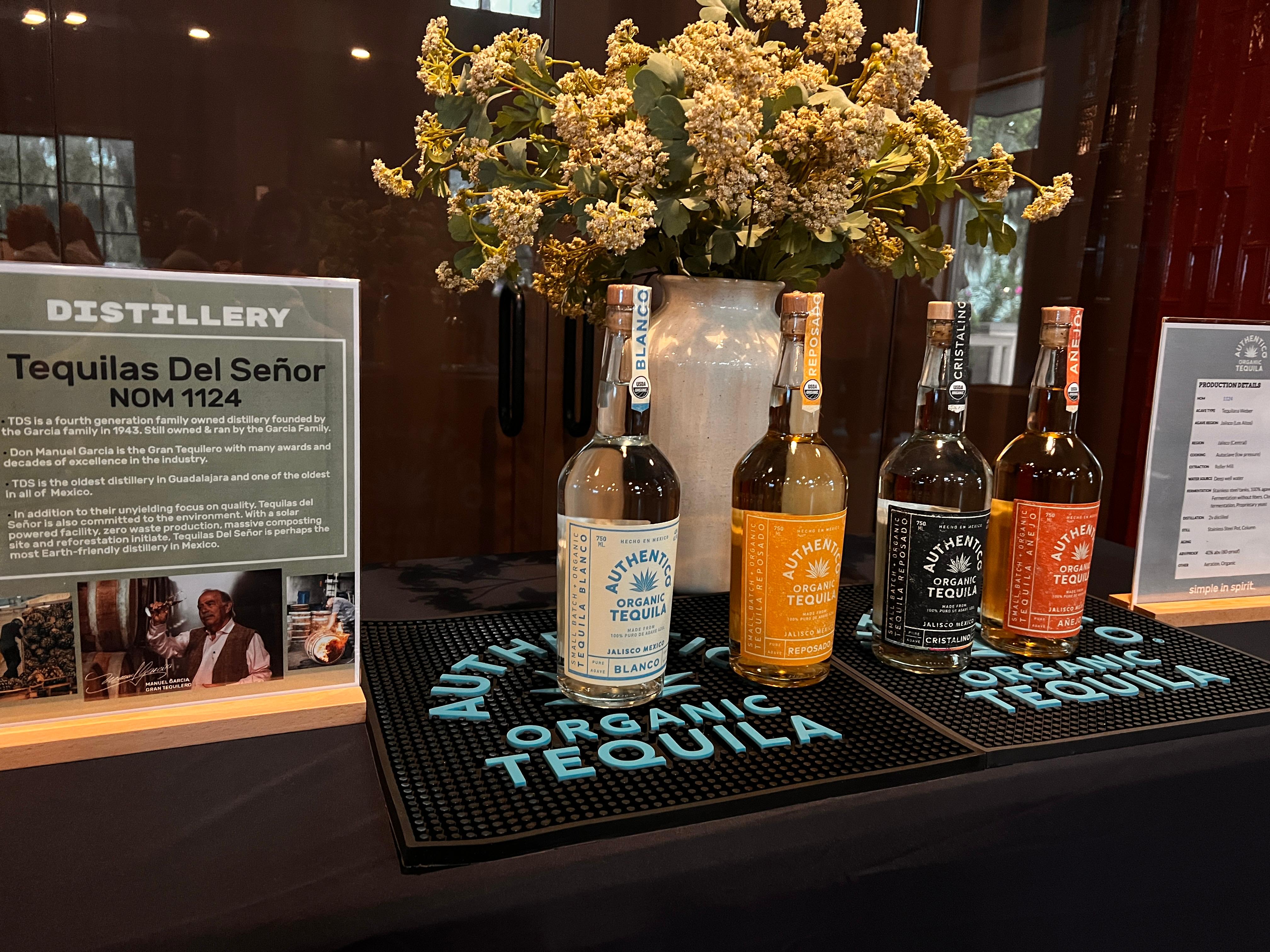 A display of Authentico Tequila featuring their Blanco, Reposado and Anejo on authenitco bar mats with flowers in the background