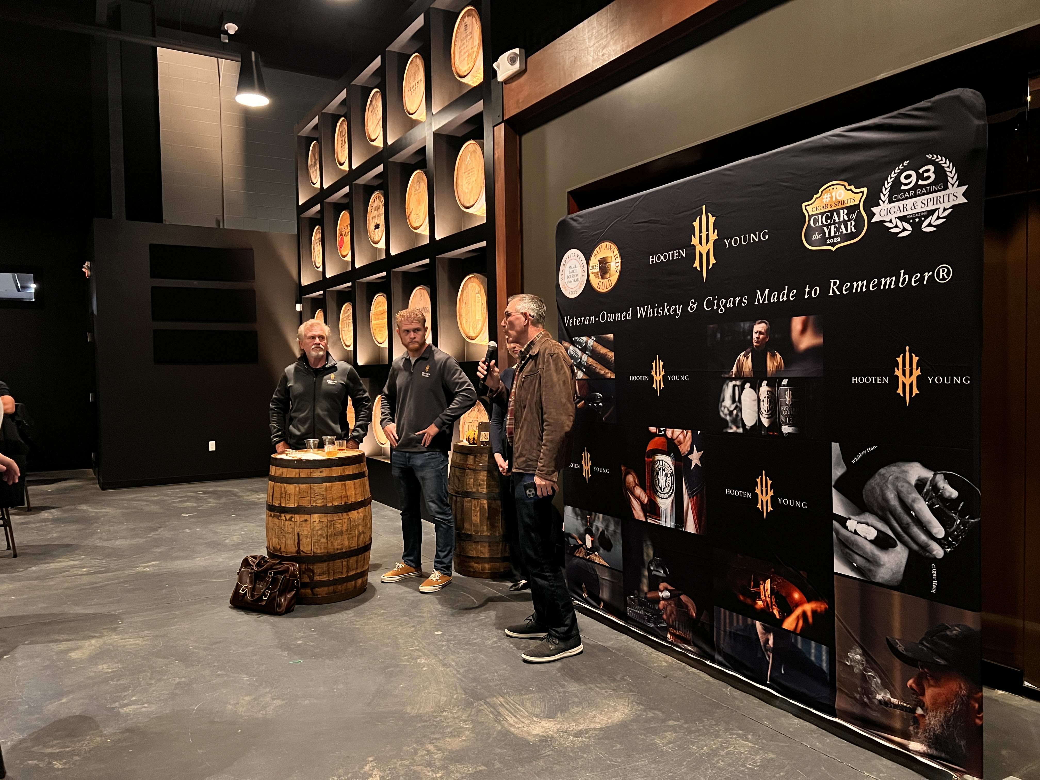 Two men standing in front of a branded poster in front of a bourbon barrel wall