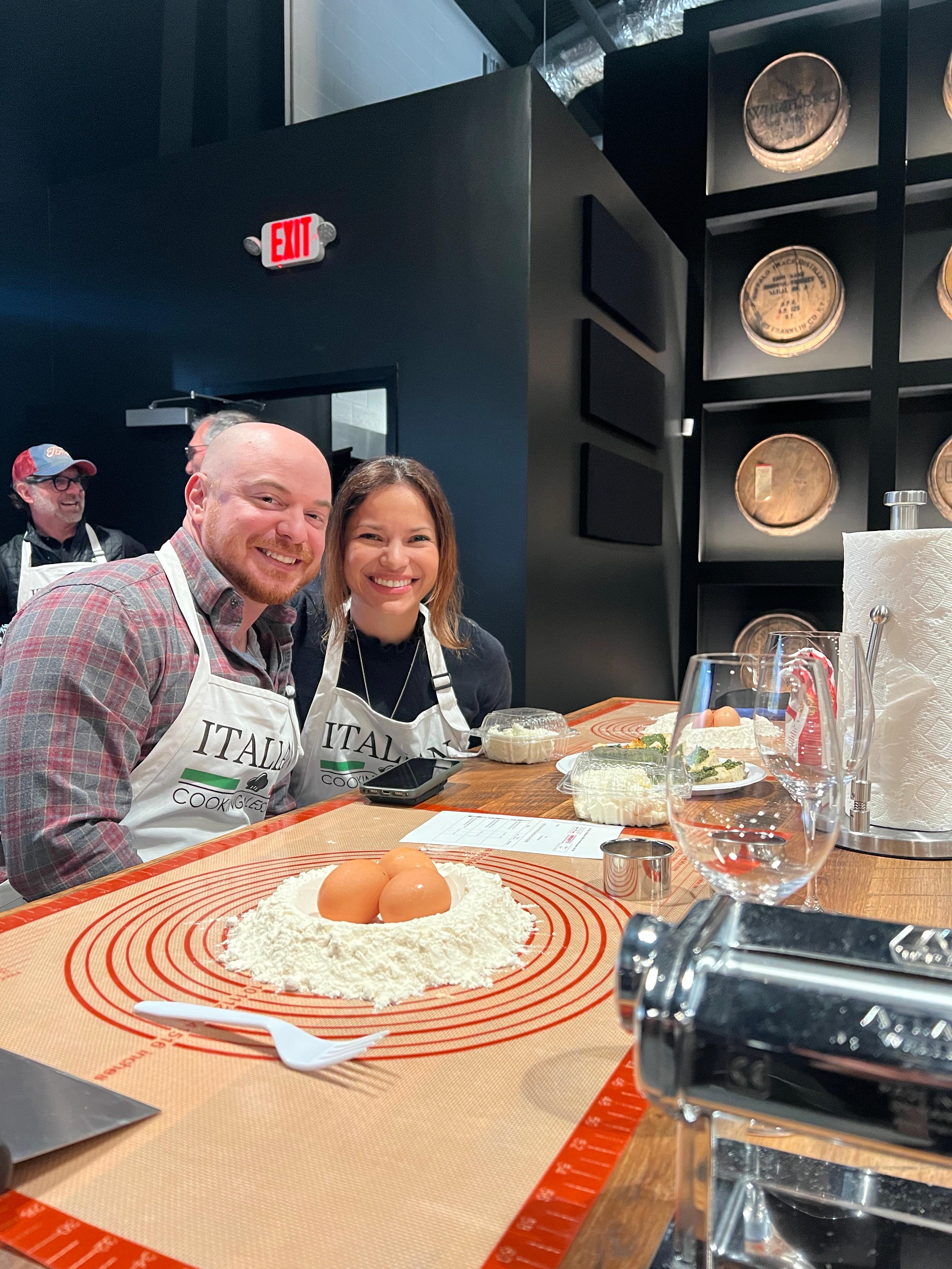 A happy couple wearing chef aprons with flour and an egg sitting on a silicone mat ready to be mixed and made into pasta