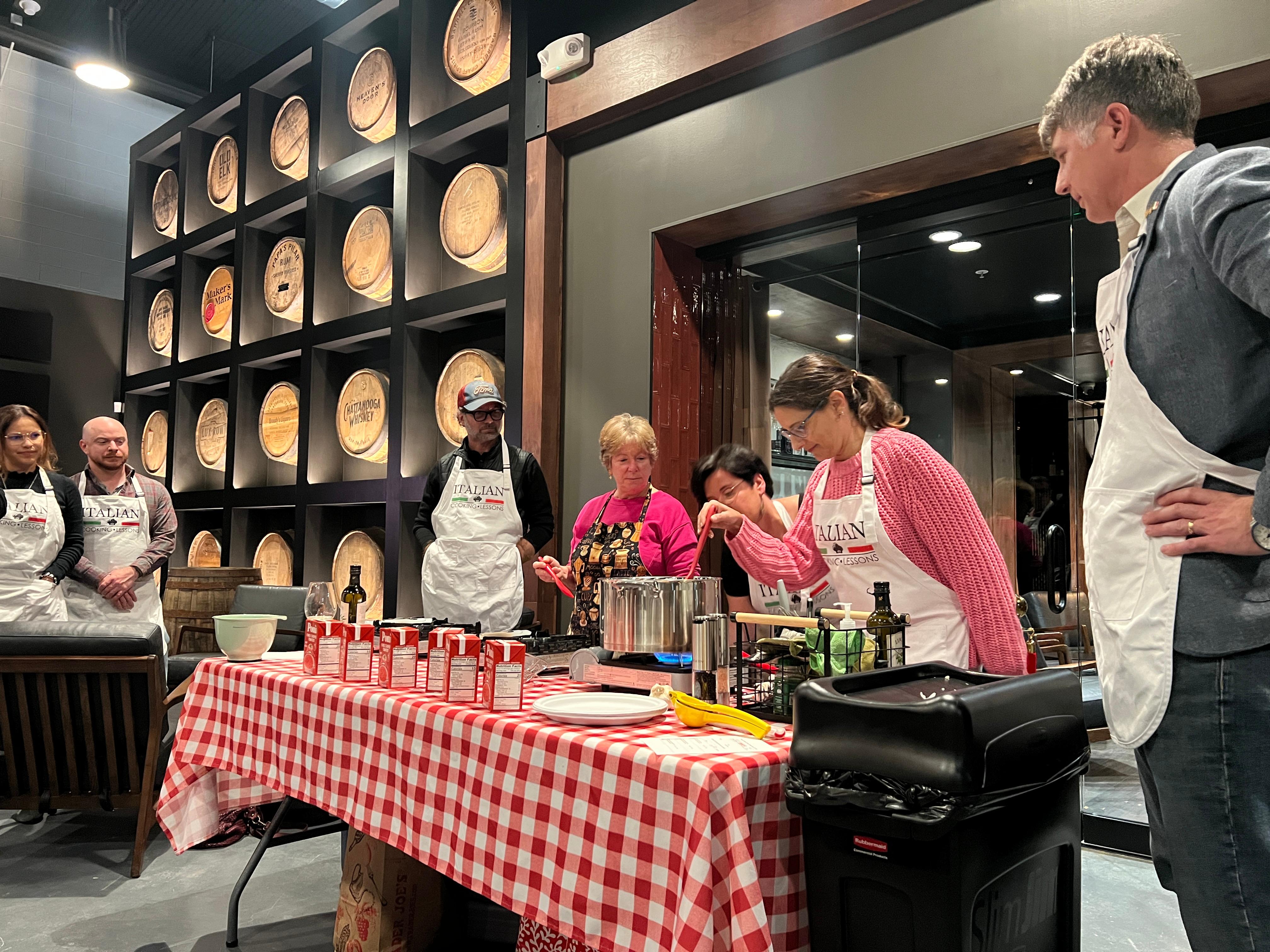 Four ladies standing around a table with a white and red table cloth with around a pot stirring homemade pasta and making red and white cheese sauce.