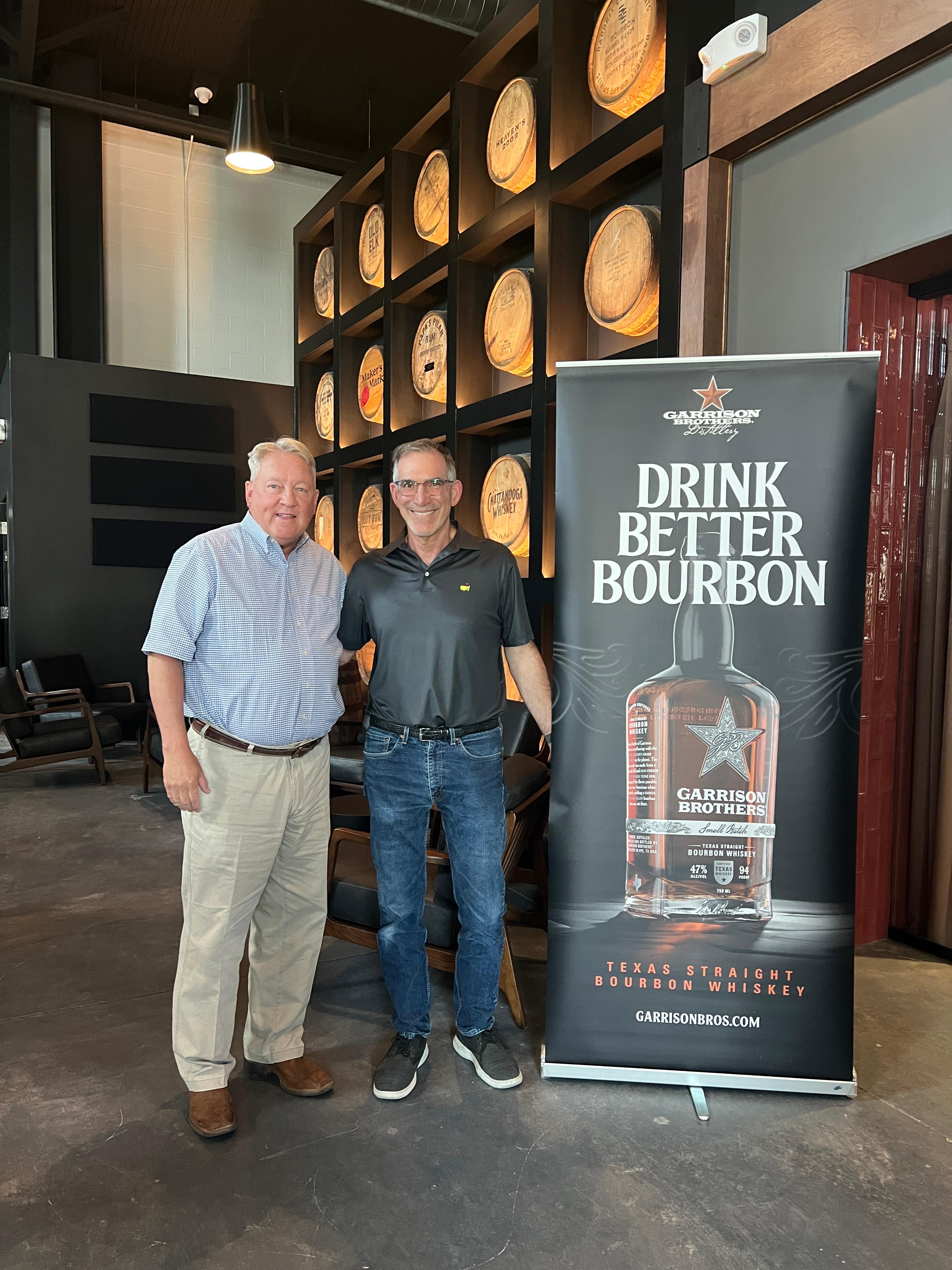 Barrel wall in the background with owner of Garrison Brothers, Dan Garrison and owner of Broudys Rickhouse, Barry Broudy standing together next to a sign that says "Drink Better Bourbon"